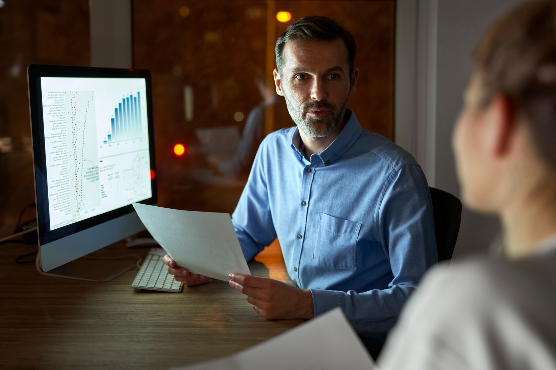 Homem de camisa azul, segurando papéis, discute dados na tela do computador com outra pessoa