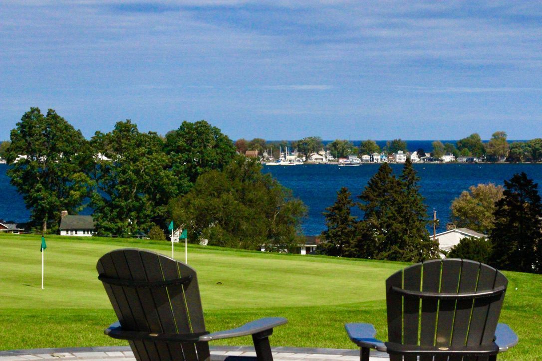 Two Adirondack chairs overlooking a green lawn, blue water, and a tree-lined shoreline under a clear sky