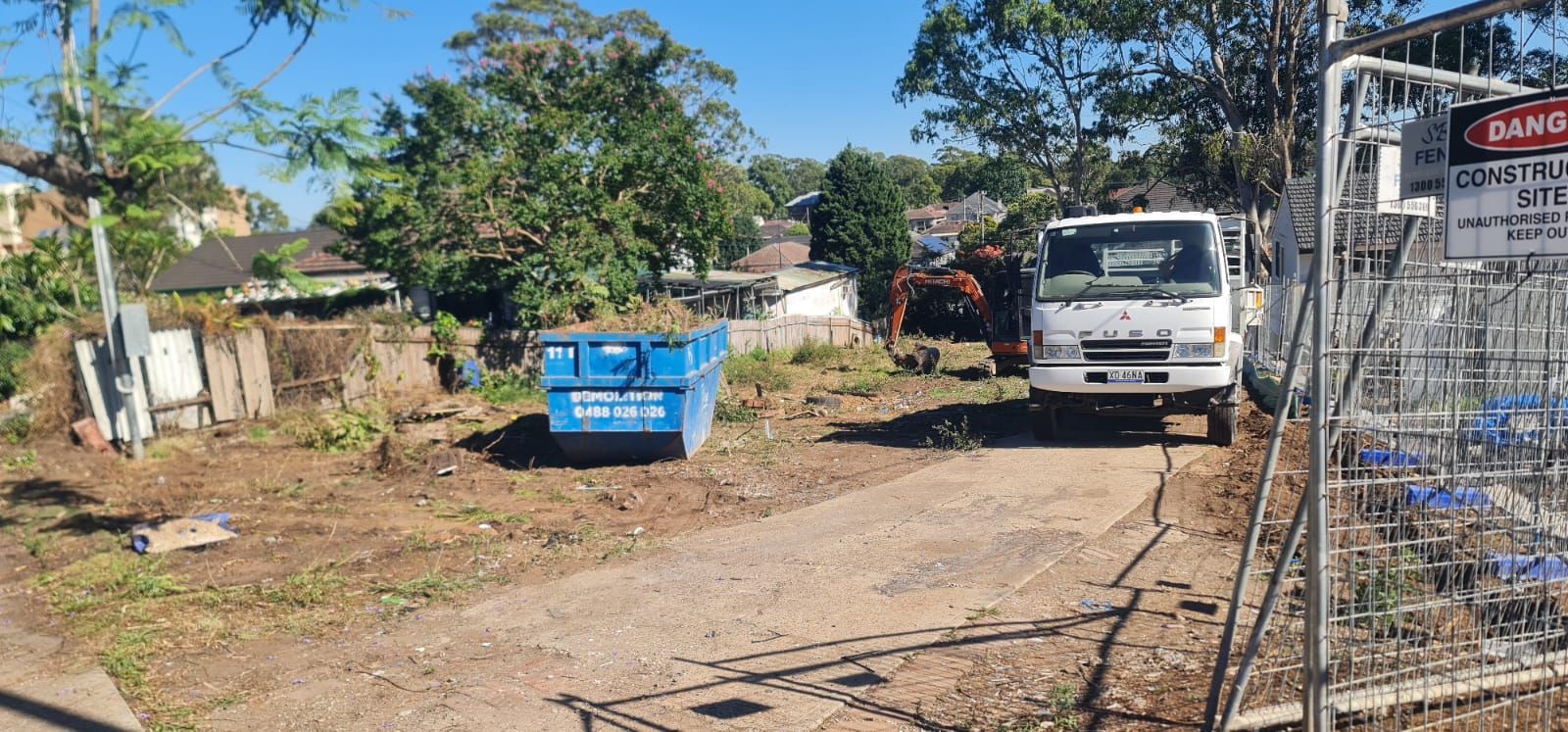 A white truck and a blue dumpster sit on a dirt lot at a construction site next to a fenced area.
