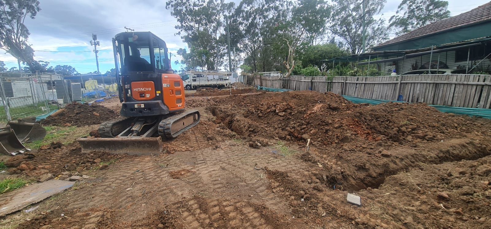 An orange excavator sits on a dirt lot in front of a wooden fence, with rows of freshly dug trenches in the foreground.