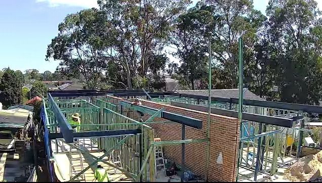 Construction site featuring a brick wall and a green steel frame for a new structure set against a background of trees.