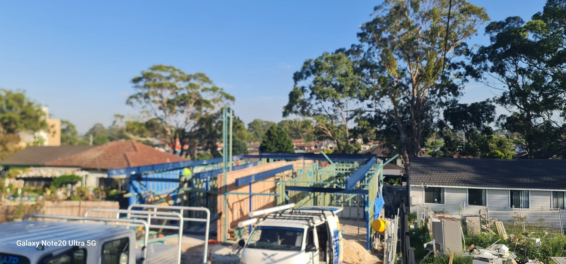 A construction site showing a blue steel frame and brick walls under a clear sky, with work vehicles in the foreground.