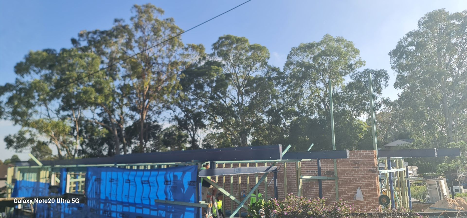 A construction site showing a brick building frame under a bright sky with tall trees in the background.