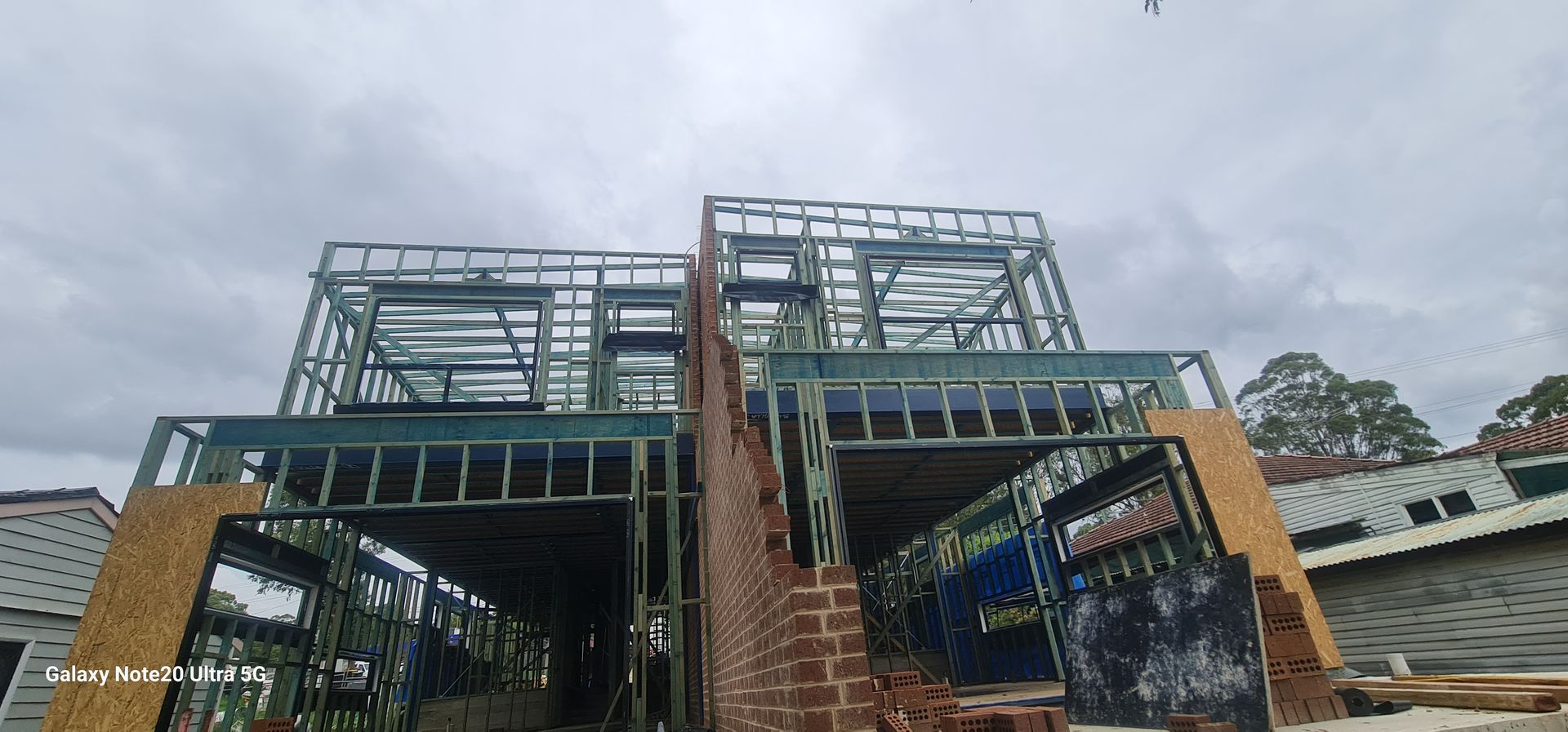 The metal frame of a two-story house under construction, partially attached to an existing brick wall under a cloudy sky.