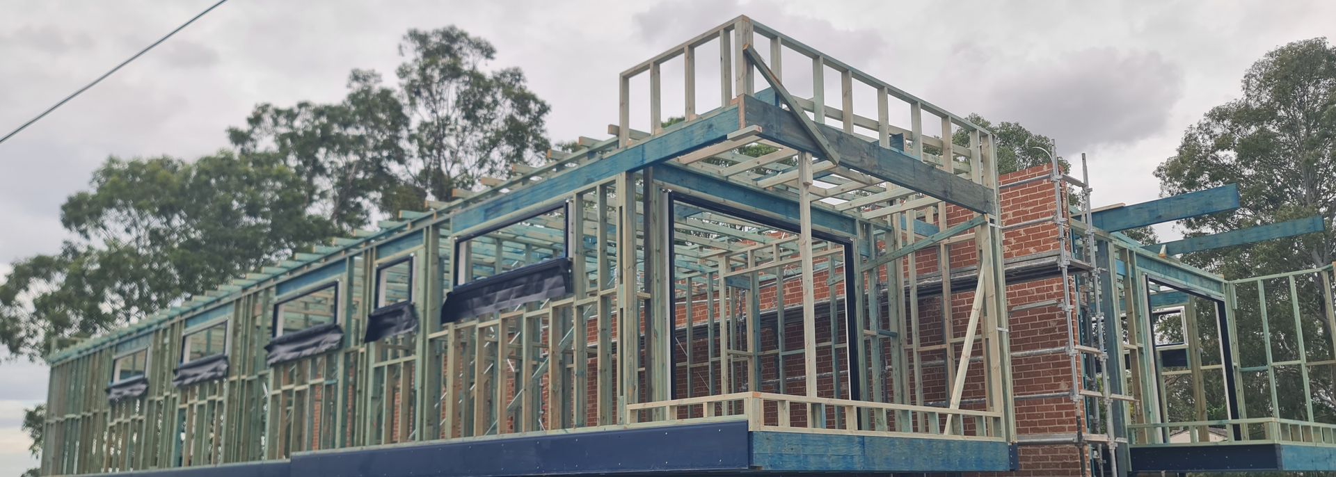 Construction site showing the metal framing of a house in progress against a cloudy sky with trees in the background.