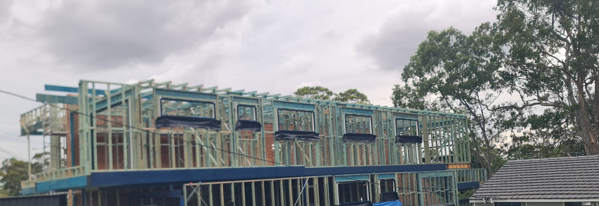 A wooden frame construction of a multi-story building against a cloudy sky with trees on the right.