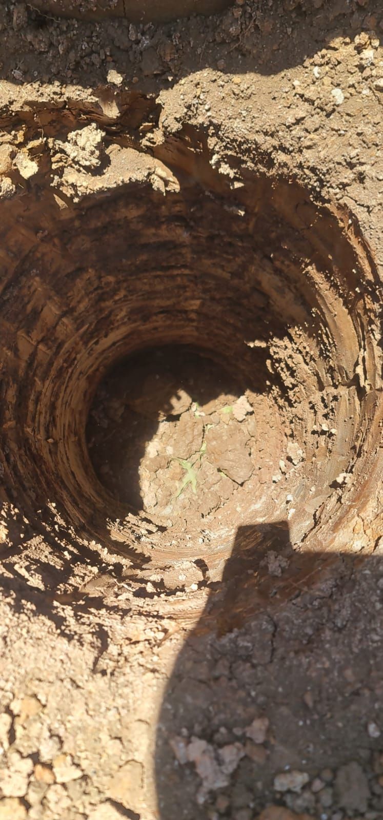 A top-down view of a deep, circular hole dug into dry, light-colored dirt, with visible drill marks on the interior walls.