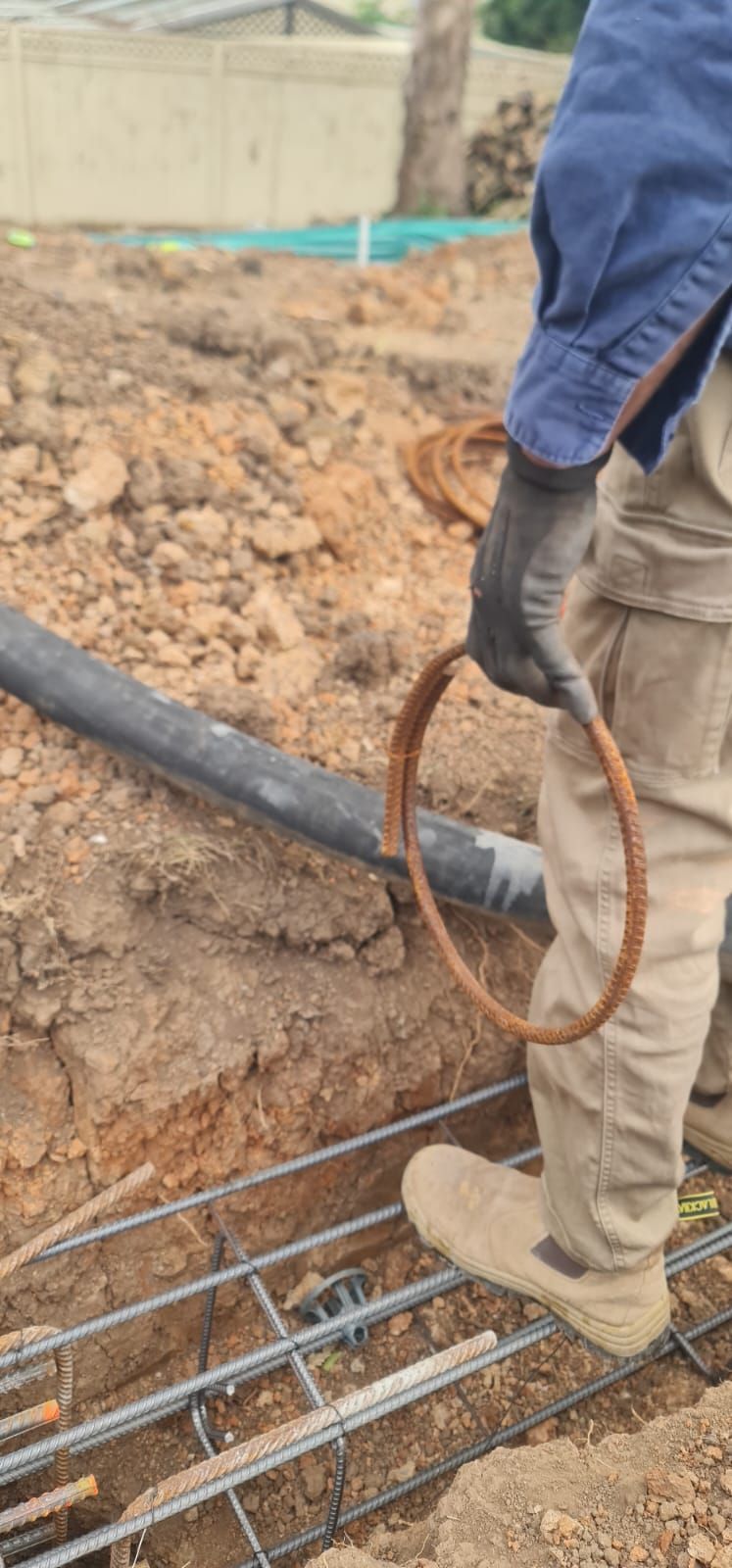 A worker holding a circular rebar hoop stands near a trench containing a structural steel reinforcement cage.