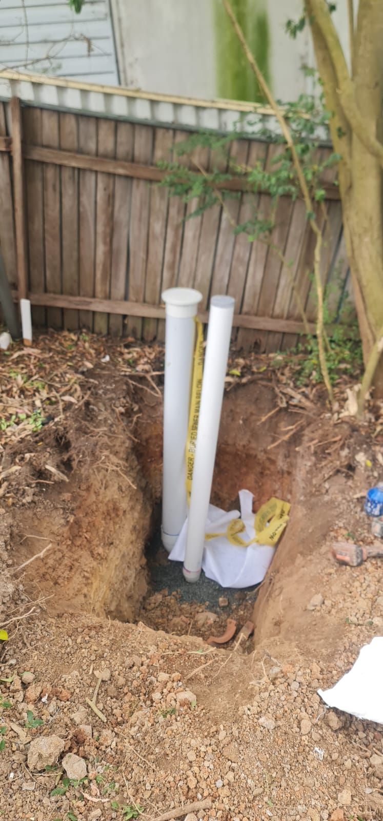 Two vertical white PVC pipes stand in a dirt excavation pit next to a wooden fence.