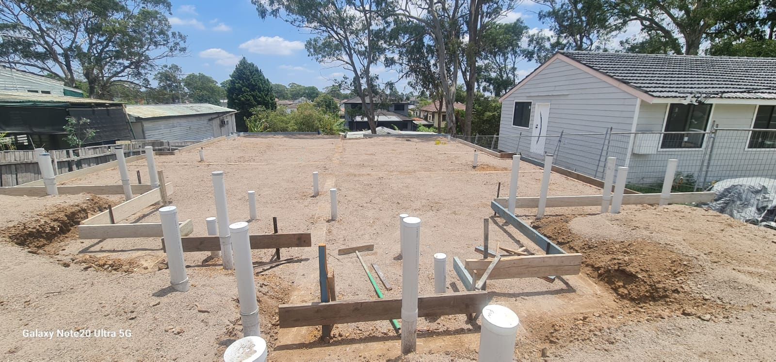 A construction site showing a prepared ground foundation with plumbing pipes and wooden formwork, near a residential house.