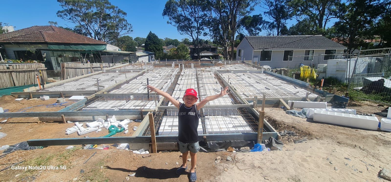 A child stands with arms outstretched in front of a residential concrete foundation under construction on a sunny day.