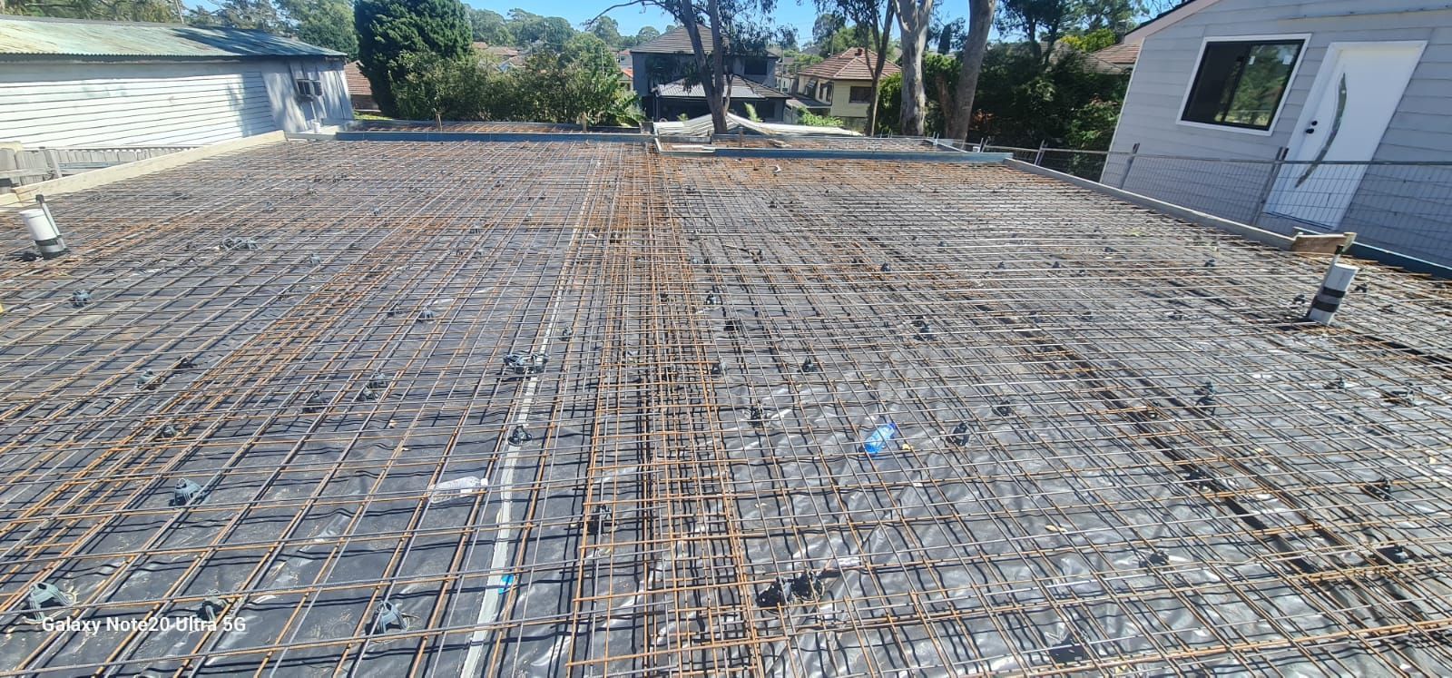 A wide-angle view of a residential construction site showing steel rebar grid framing over a foundation slab.