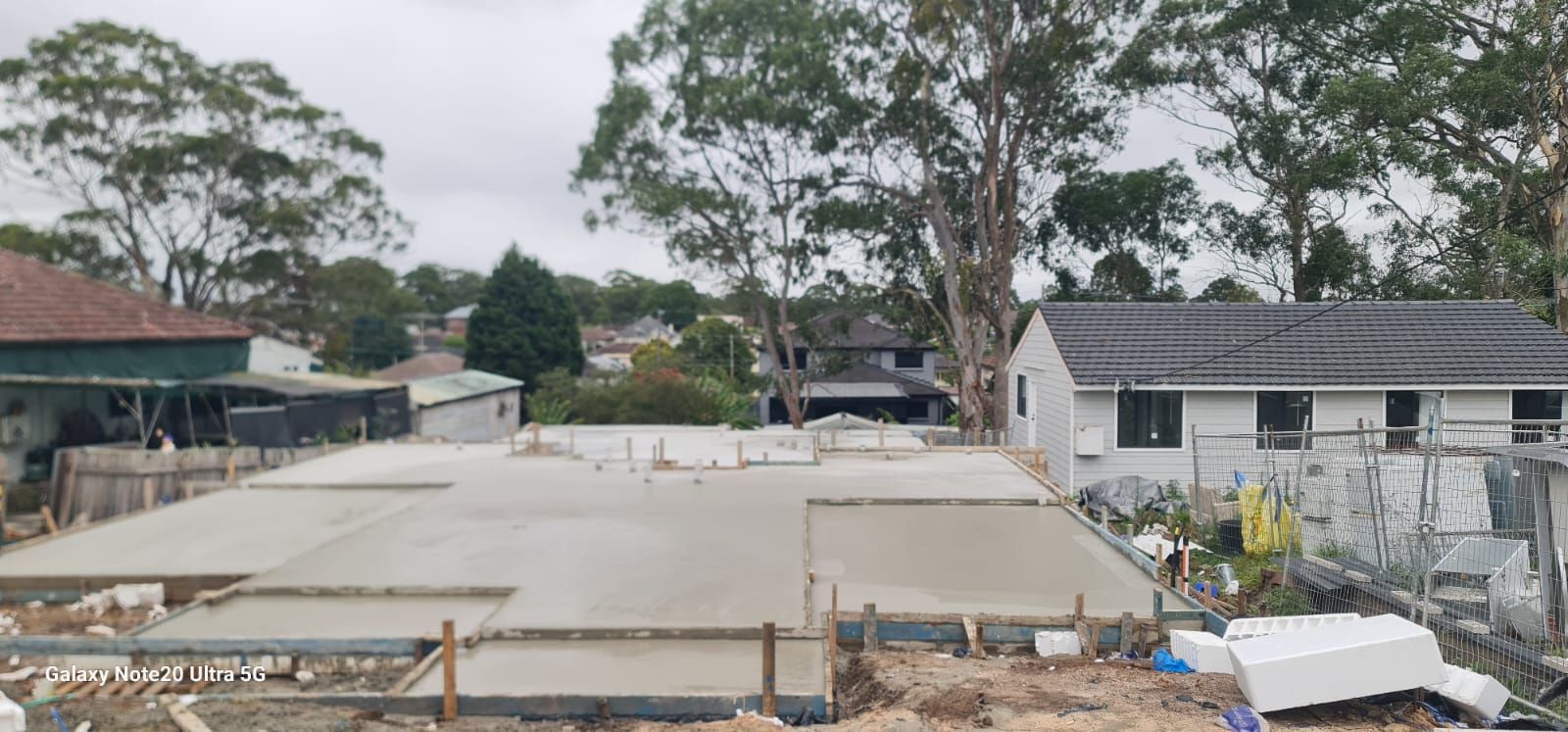 A freshly poured concrete slab foundation for a new house build on an overcast day, with neighboring homes in the background.