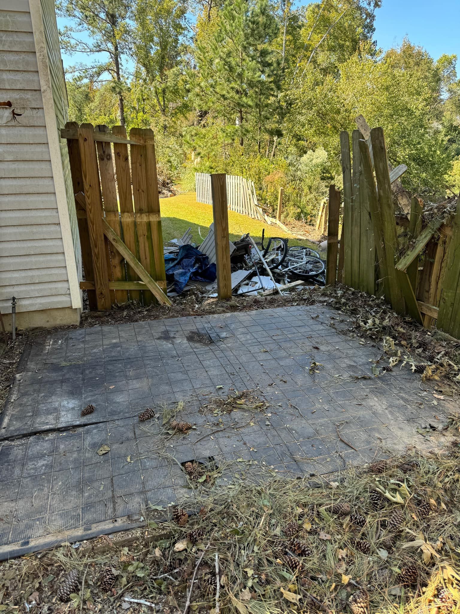 Patio with a damaged wooden fence, debris, and surrounding trees.