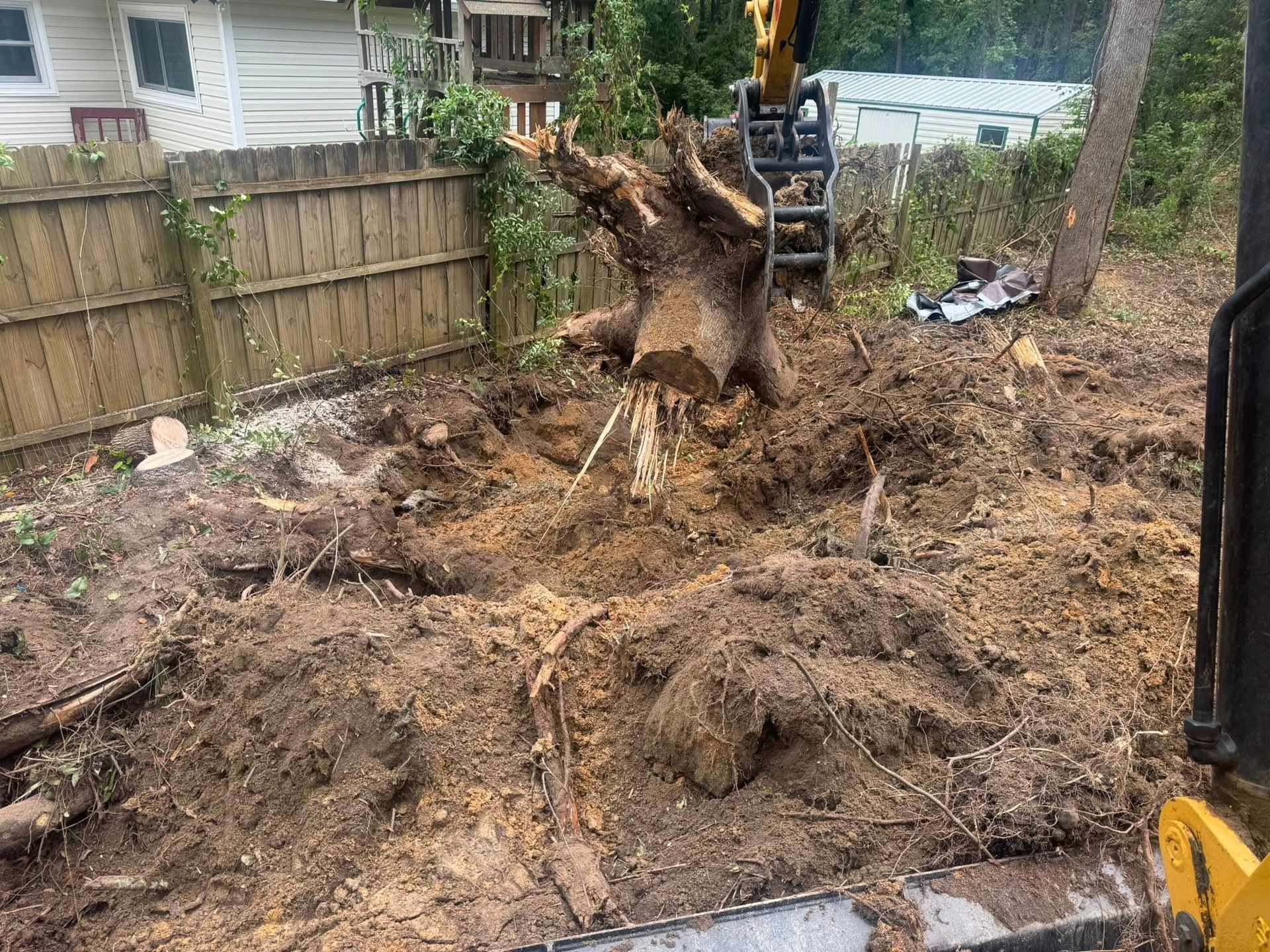 Excavator removing a tree stump in a backyard. Earth and wood debris surrounds the stump near a wooden fence.