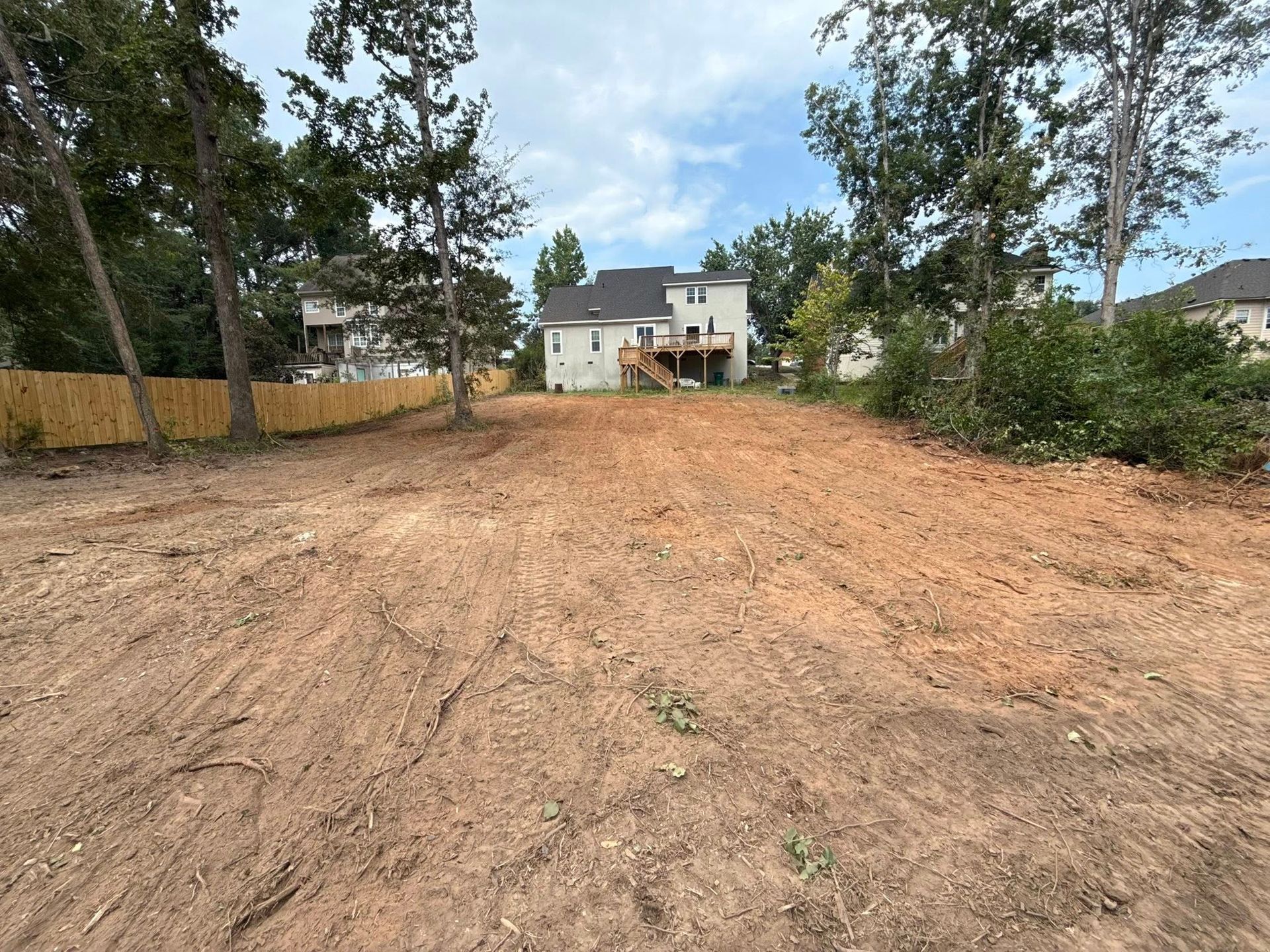 Cleared dirt lot, bare with tire tracks, backing onto a two-story house with trees on either side.