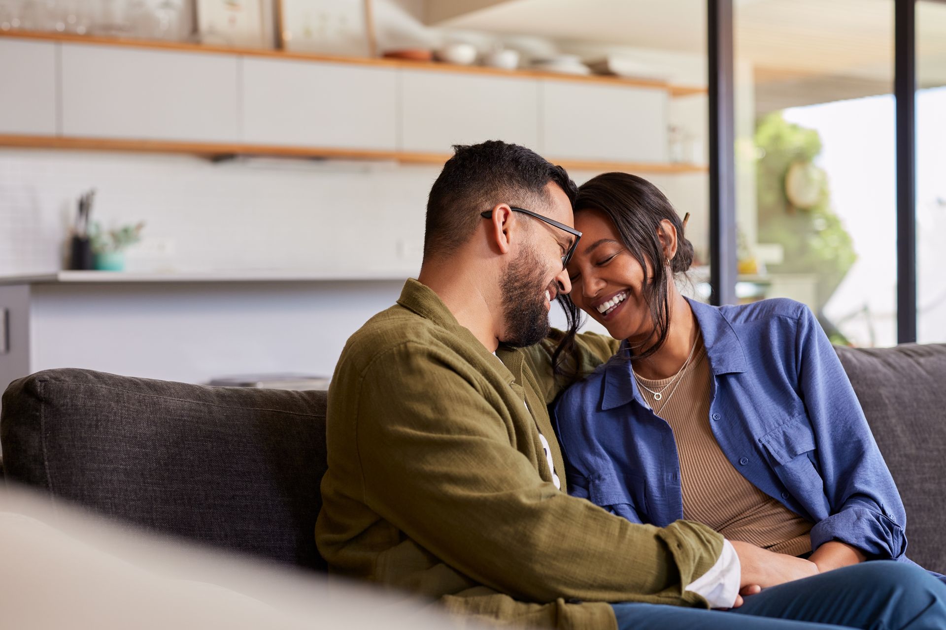 A couple smiling and leaning their heads together while sitting on a sofa in a modern home living room.