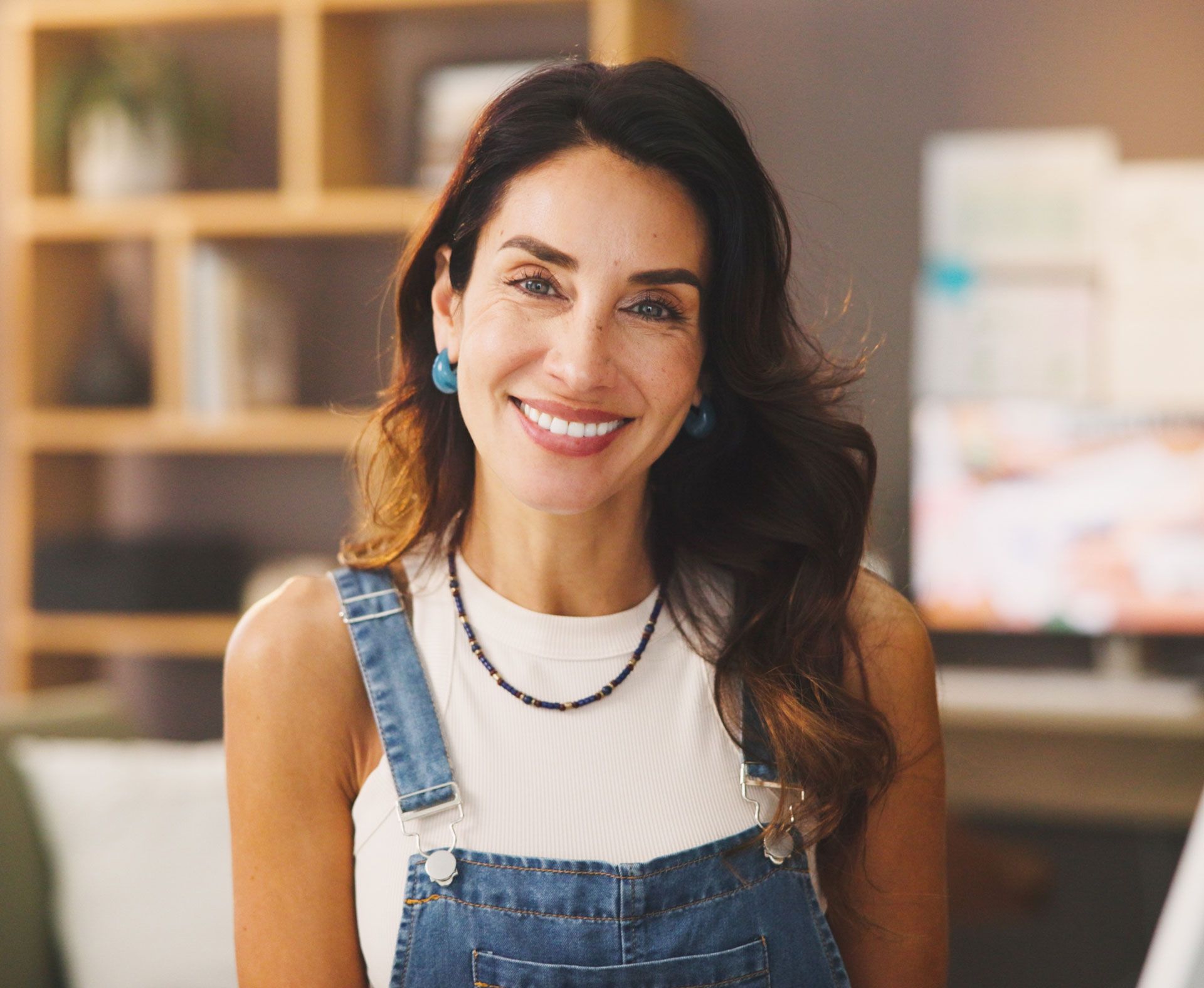 Smiling person in blue denim overalls and a white top, positioned in a blurred indoor office or home workspace.