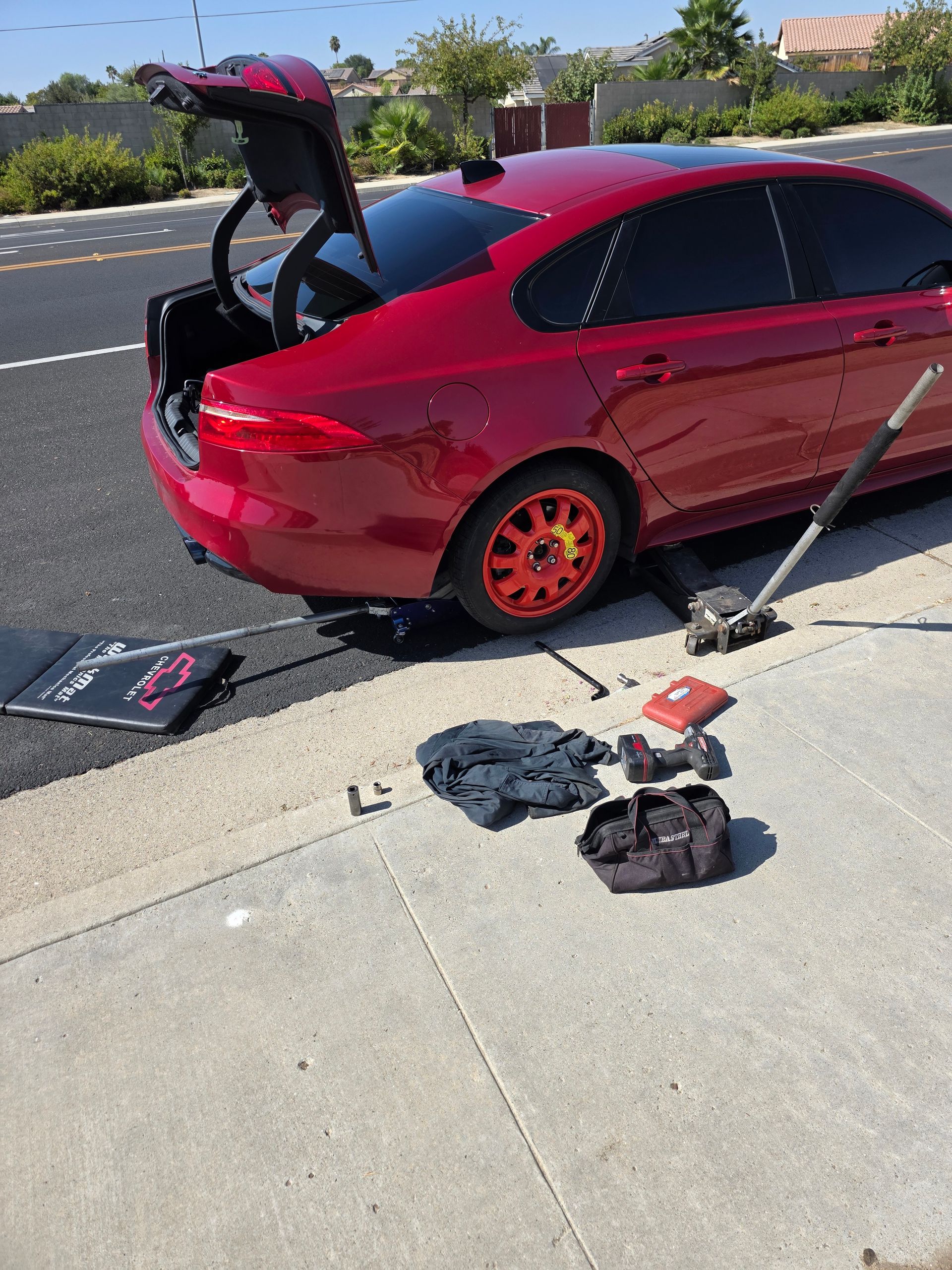 Red car with orange wheels being worked on with tools on a sidewalk next to a street.