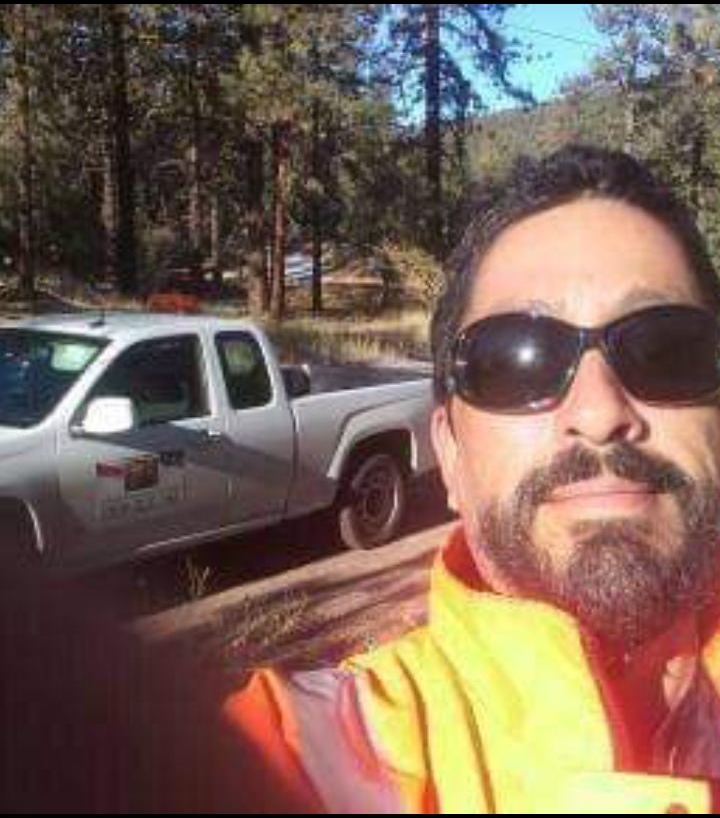 Man in sunglasses and orange vest with truck in wooded area.