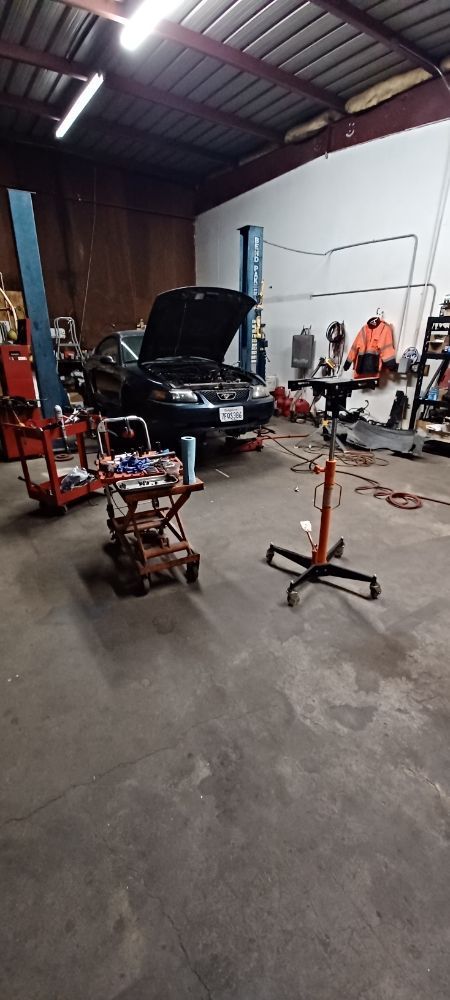 A car repair shop. A dark-colored car is under repair with its hood up. Tools and equipment are around.