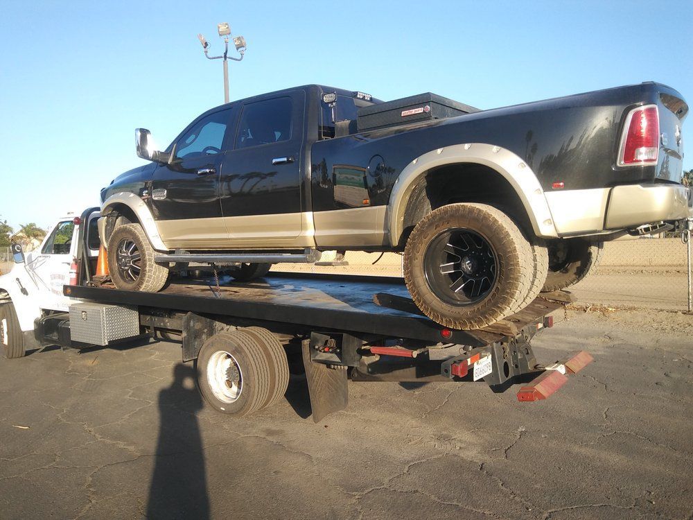 Black and tan pickup truck being towed on a flatbed tow truck on a sunny day.