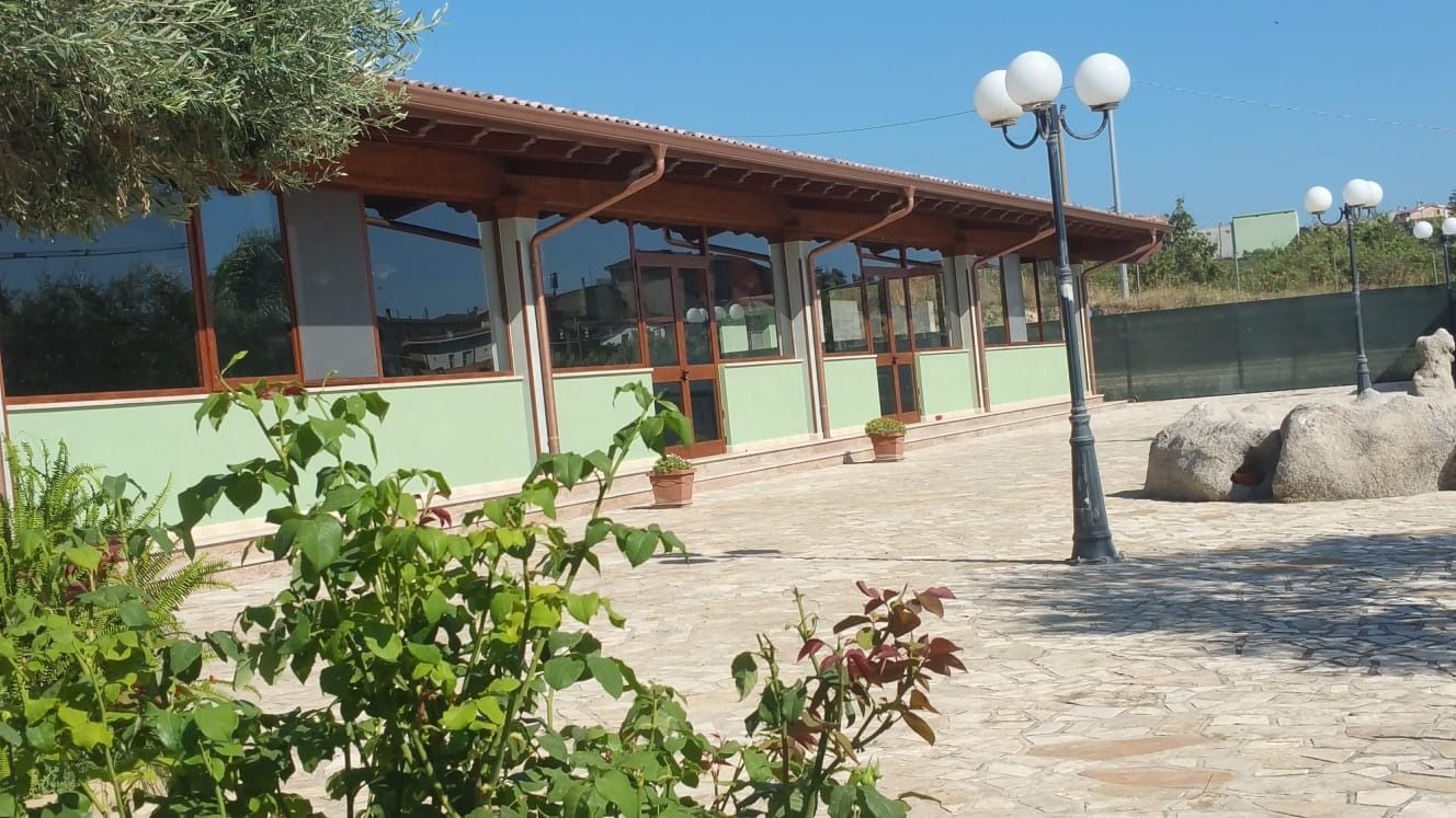 Ristorante con ampie finestre, esterno verde chiaro e cortile con ghiaia sotto un cielo azzurro brillante.