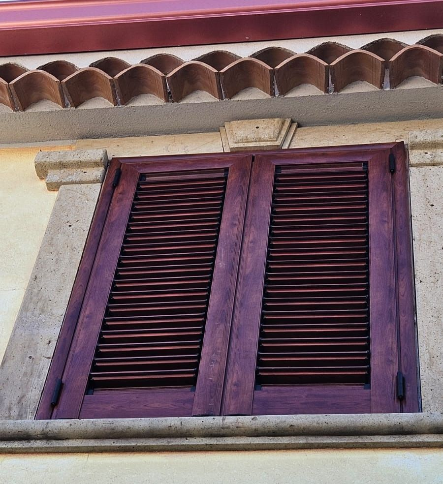 Persiane in legno viola su una finestra con finiture in pietra beige, sotto un tetto in tegole di terracotta.