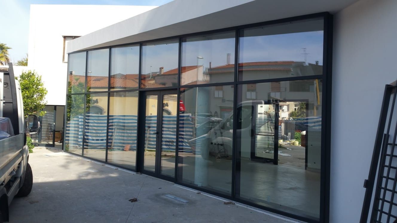 Un edificio moderno con una grande facciata in vetro riflette l'ambiente circostante. Infissi neri, pareti bianche e cielo azzurro.