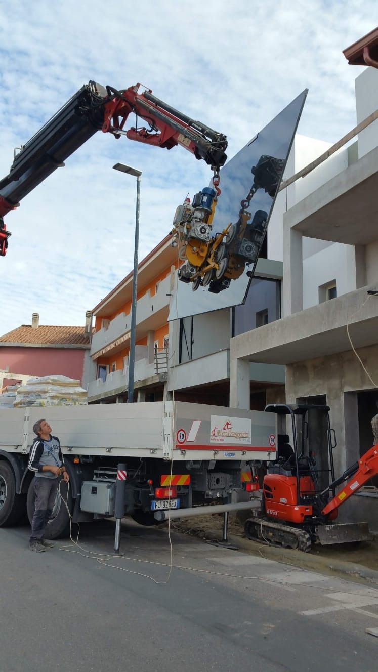 Una gru solleva un grande oggetto a specchio sopra un camion in una strada piena di edifici. Un uomo osserva.