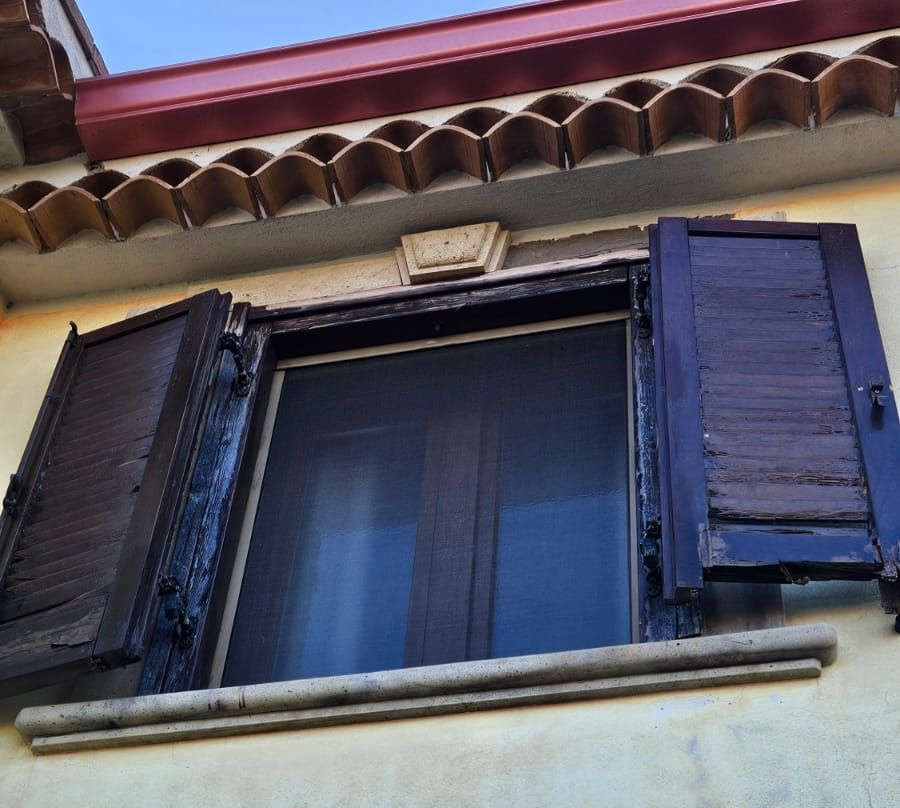Finestra con persiane in legno aperte e consumate dal tempo su un edificio giallo. Tetto con tegole marroni.