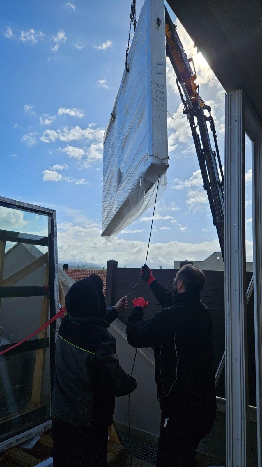 Due operai guidano un grande pannello sospeso da una gru contro un edificio in una giornata di sole.