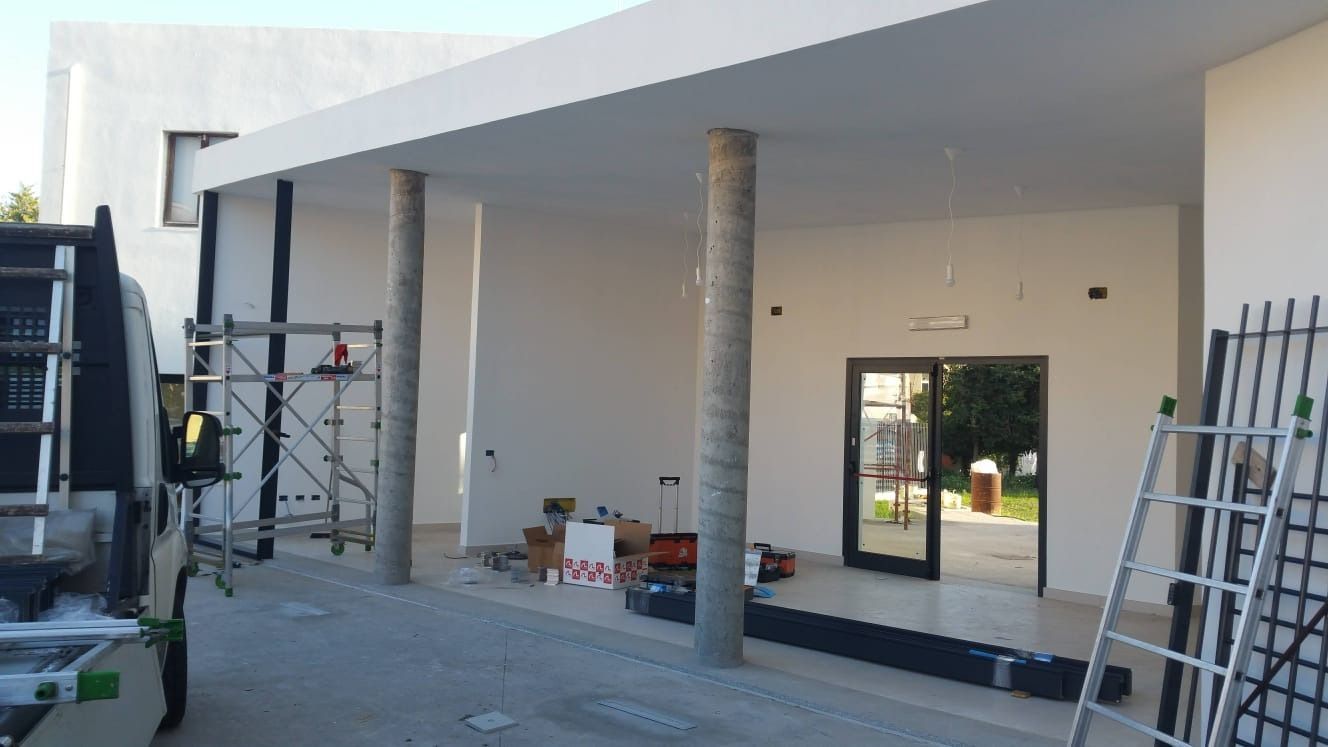 Esterno di un edificio in costruzione, con colonne, pareti bianche e una porta visibile.