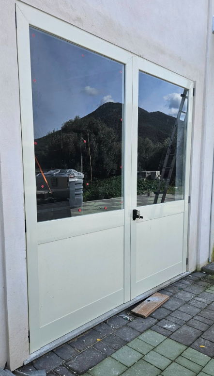 Doppie porte bianche con grandi pannelli di vetro, che riflettono una montagna e il cielo, incastonate in un edificio dai colori chiari.
