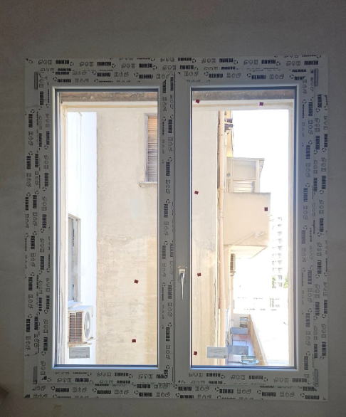 Doppia finestra con cornice bianca che si affaccia su un paesaggio urbano; la luce del sole illumina la vista.