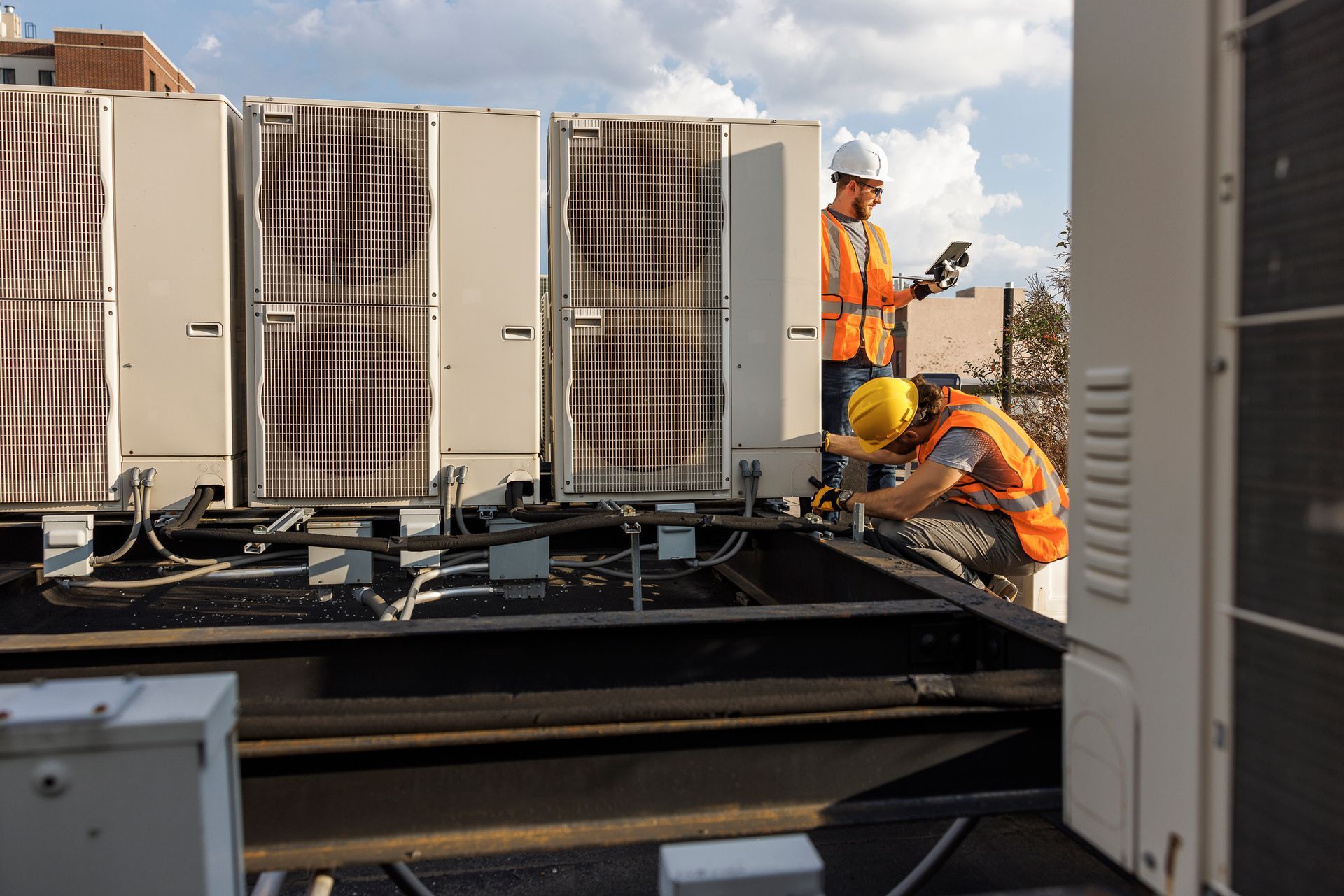 Two HVAC technicians in safety gear inspect rooftop commercial air conditioning units.