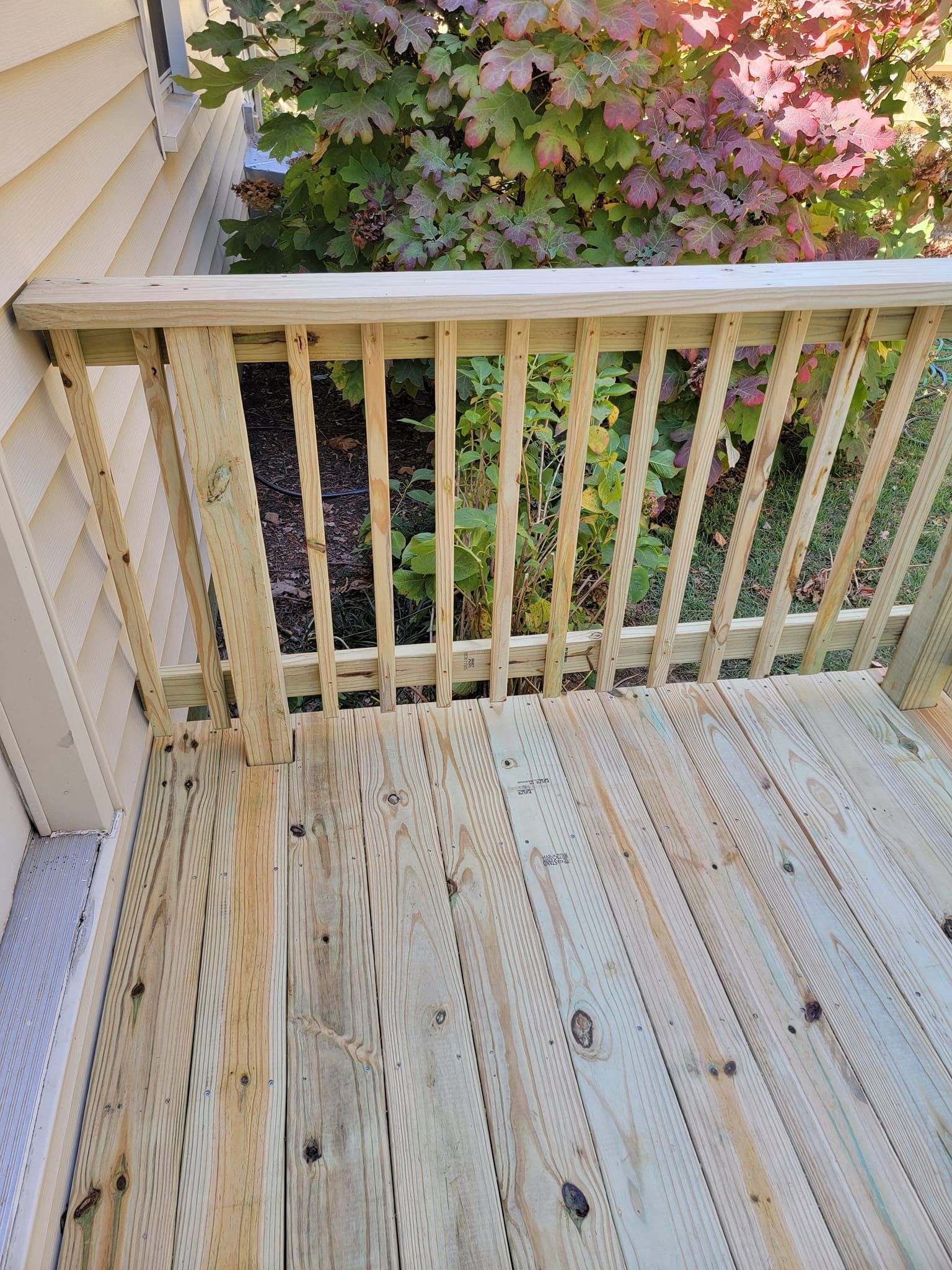 Wooden deck with railing; yard with green and red foliage in the background.