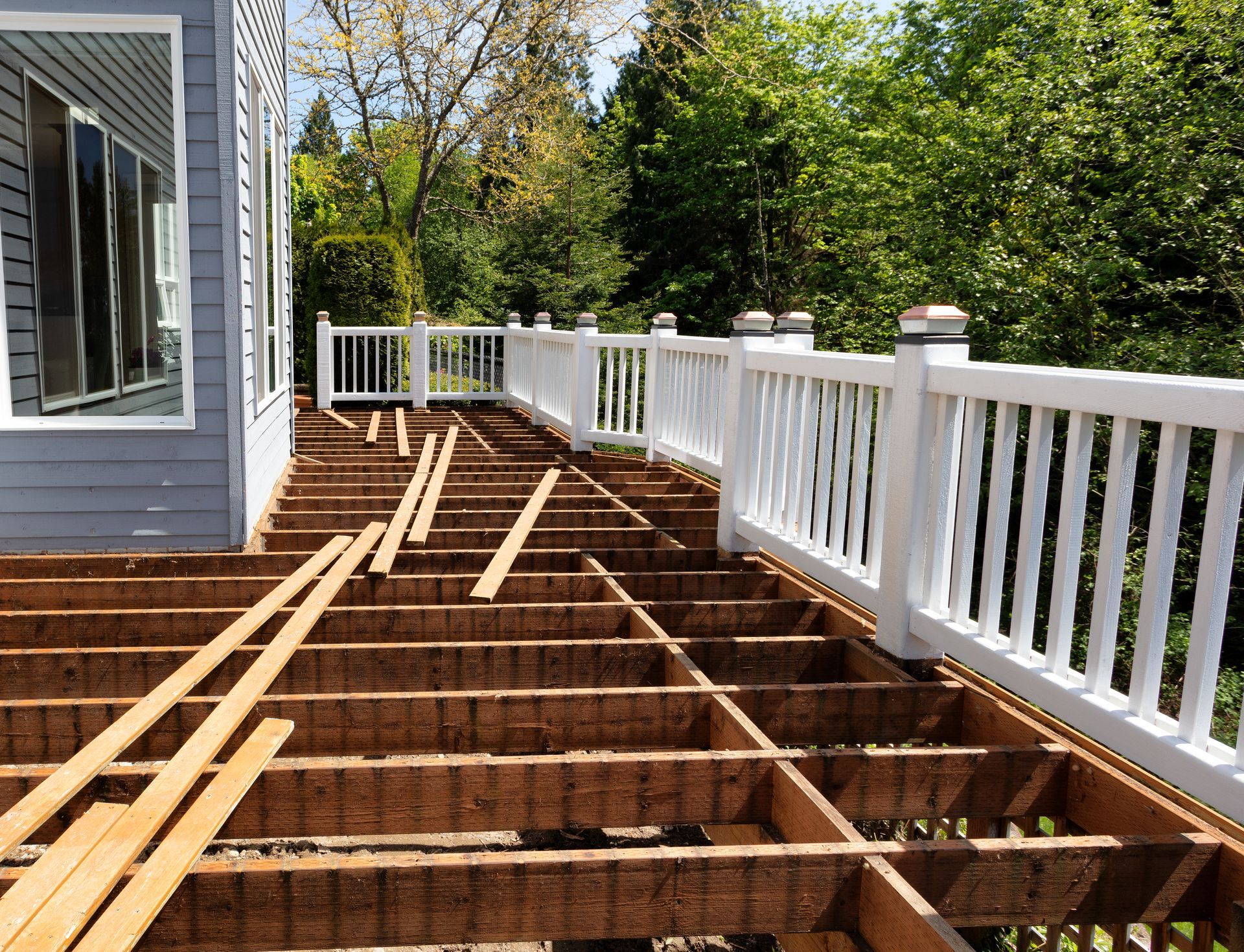 Deck in the process of being repaired, showing exposed beams and white railing along the edge; house and trees in background.