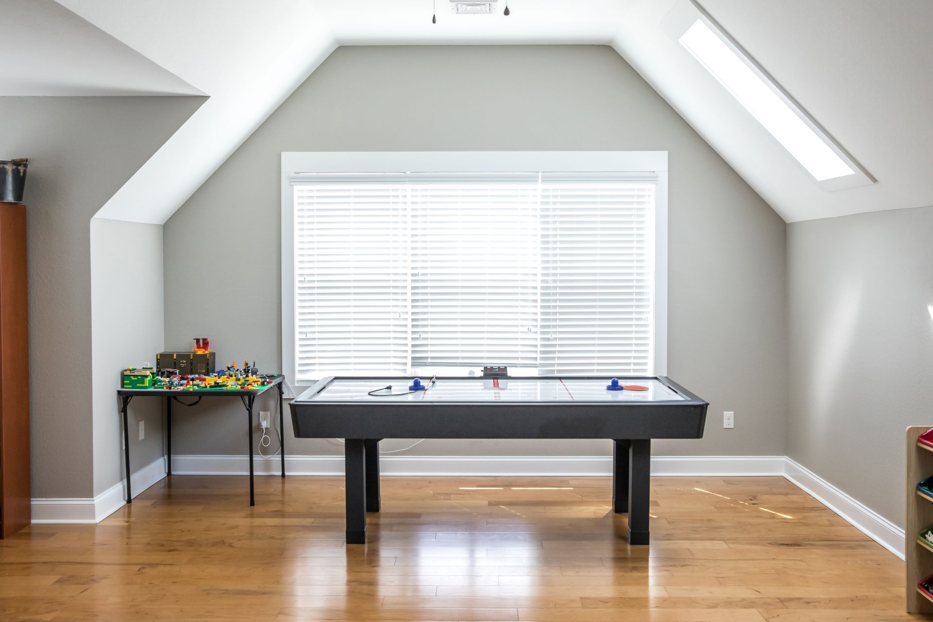 Playroom with air hockey table, small Lego table, and large window with blinds. Hardwood floors, gray walls.