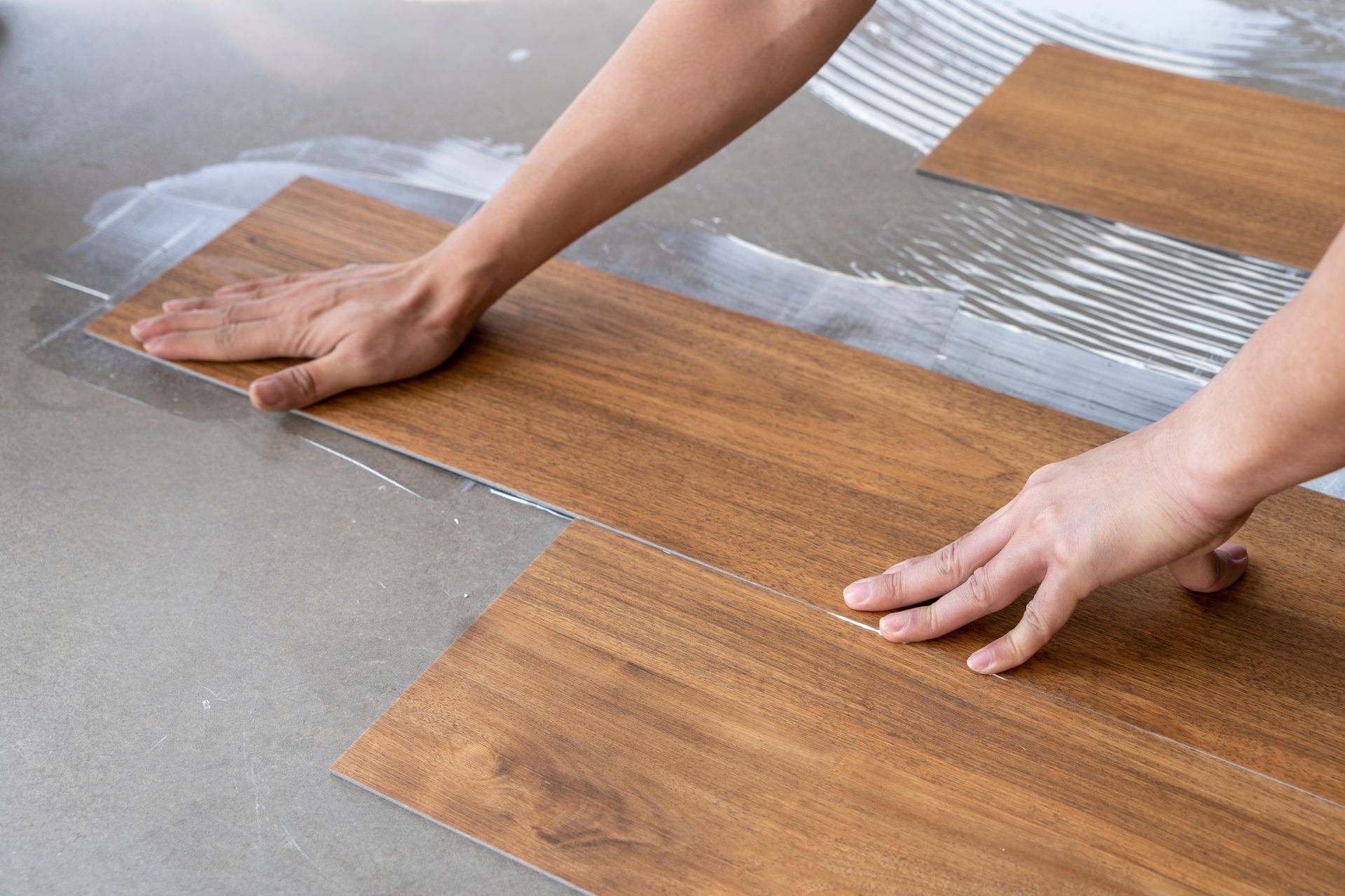 Hands installing wood-look flooring planks on a concrete surface, using adhesive.