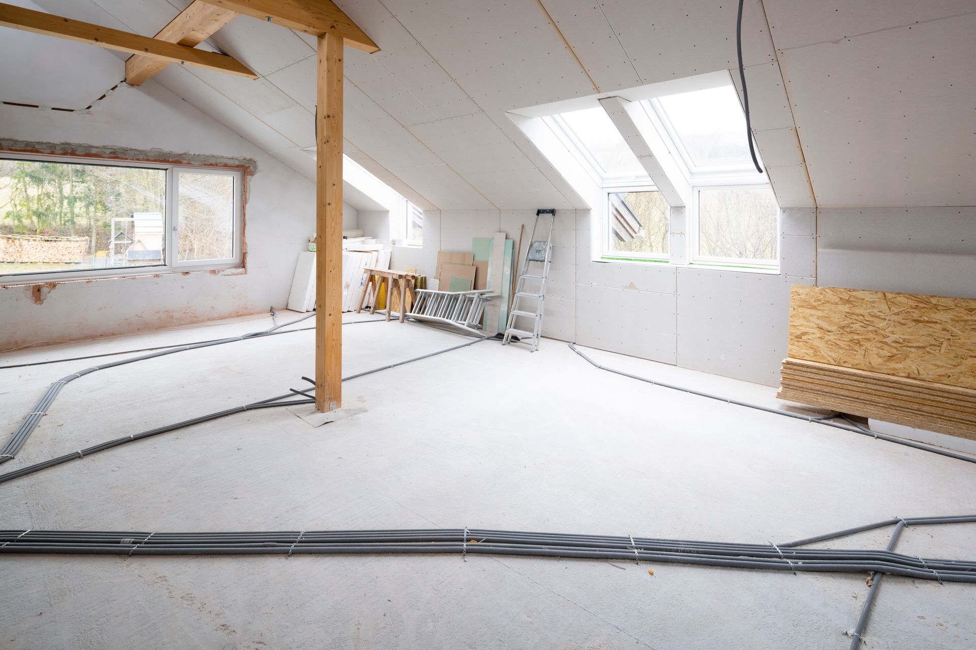 An unfinished attic room with exposed wiring, drywall, windows, and wooden support beams.