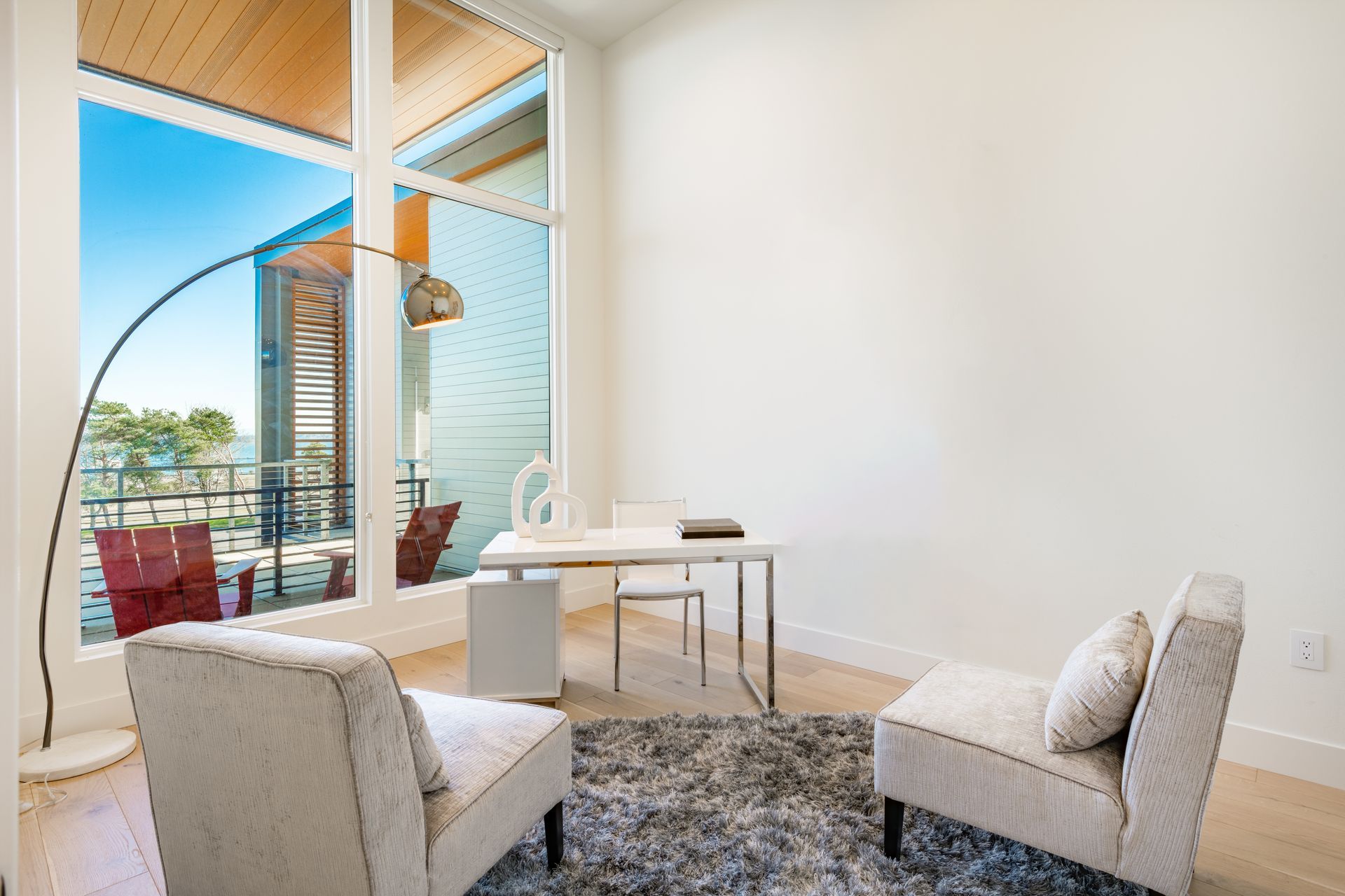 Office space with large window overlooking a balcony. White desk, two chairs, rug, and floor lamp.