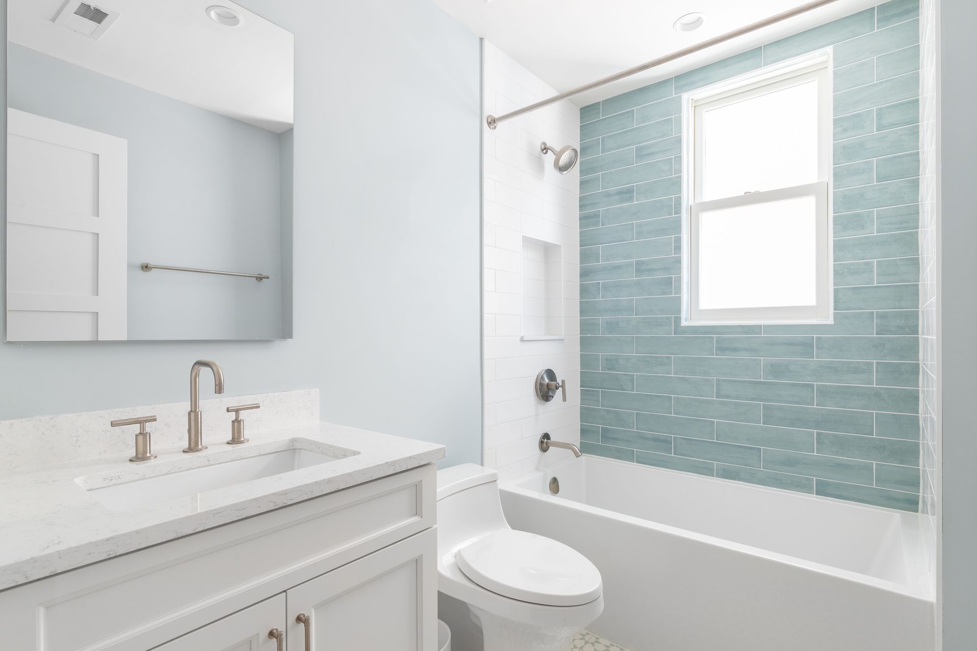 A modern bathroom with light blue walls, white vanity, toilet, and bathtub; teal tile backsplash.