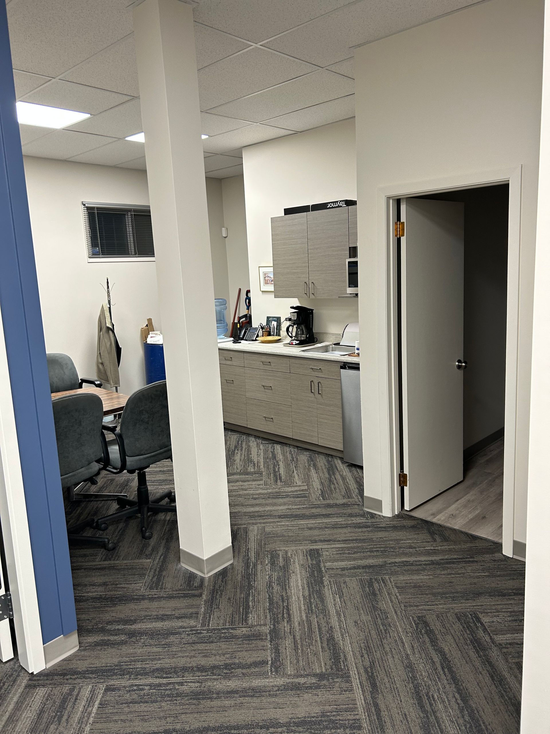 A room with a desk and chairs in it and a kitchen in the background.