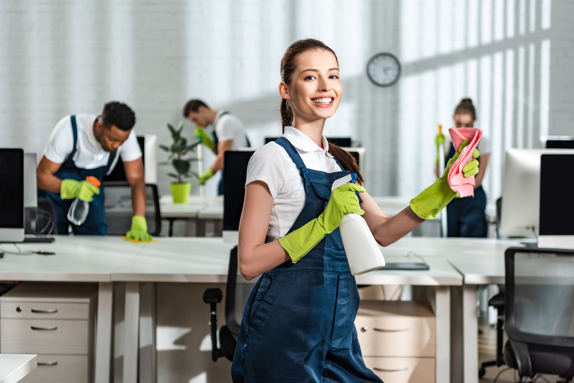 Office cleaners smiling, wiping desks, wearing gloves and overalls.