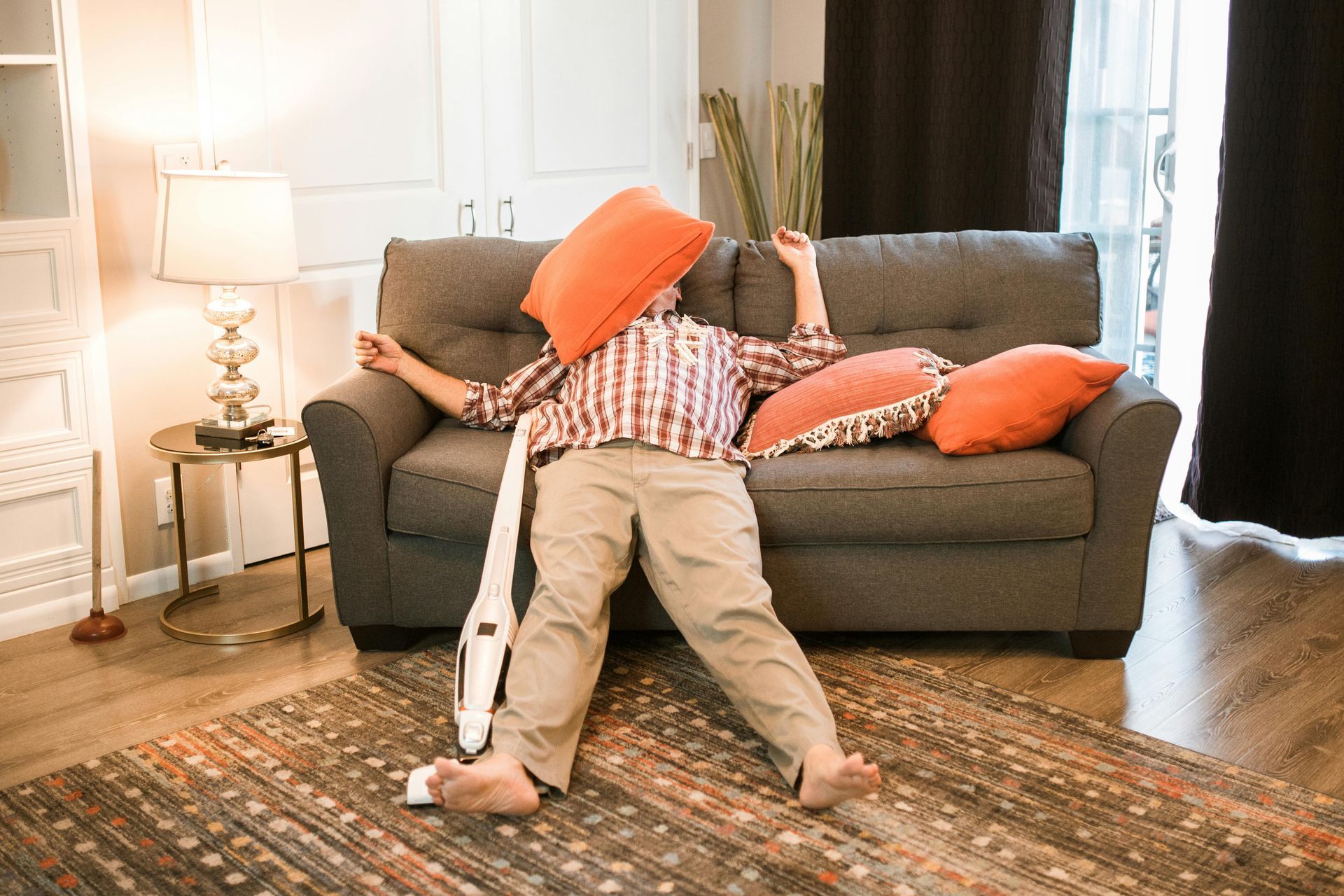 A person lying on a couch with an orange pillow over their face, holding a white handheld vacuum.