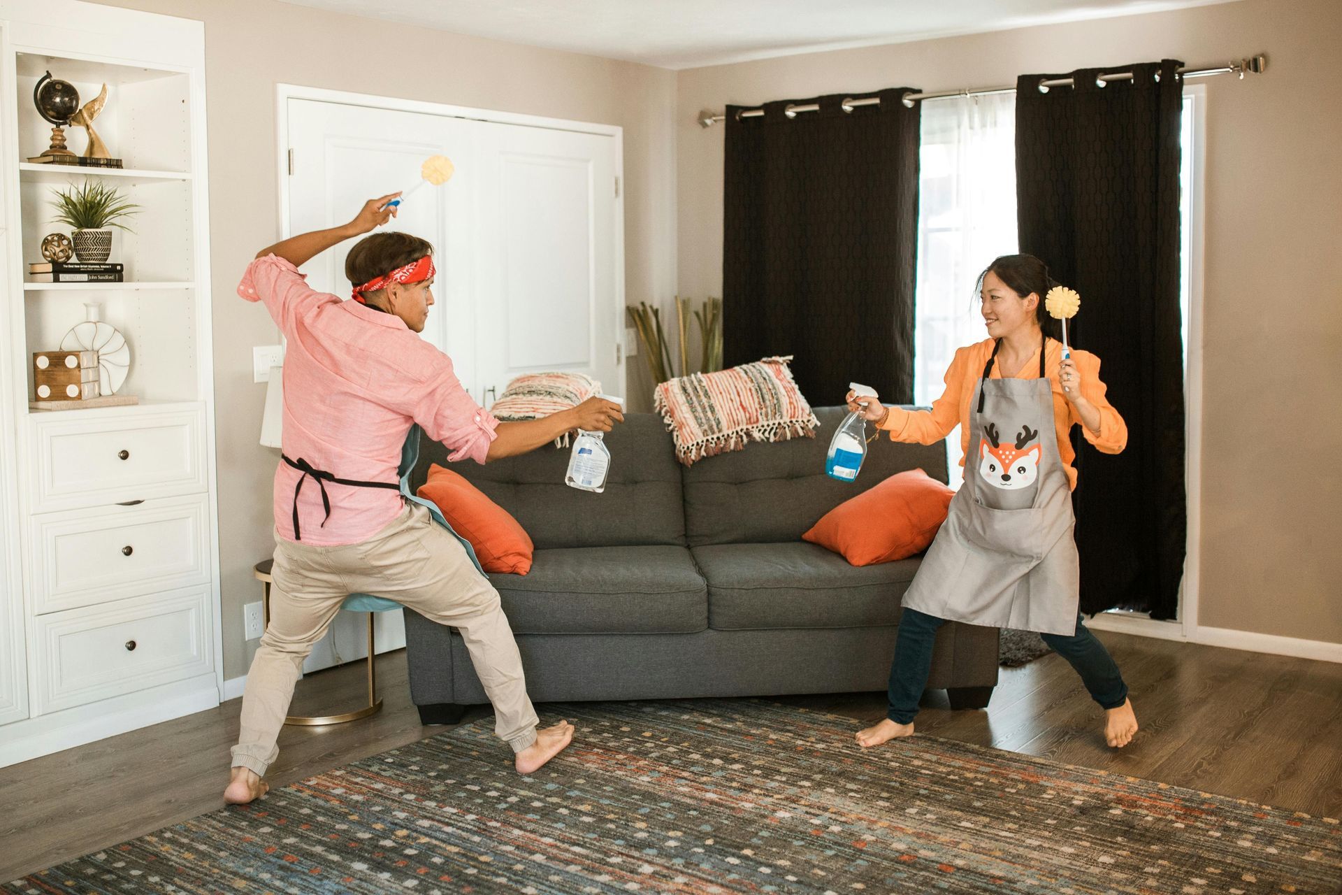 Two people in aprons playfully pose as if dueling with a spray bottle and cleaning brush in a living room.