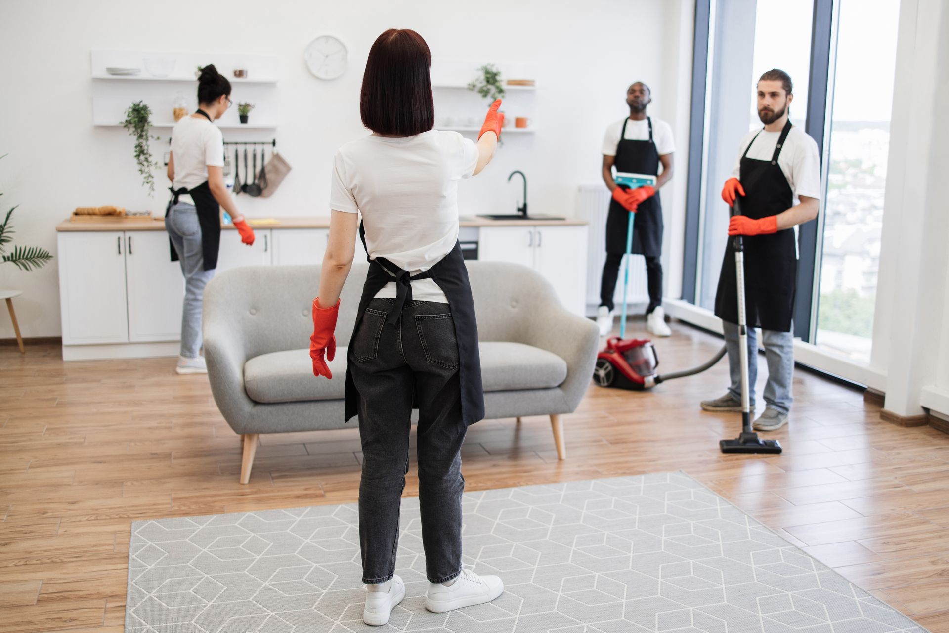 Cleaning crew in aprons sanitizing a modern apartment. One points, others vacuum, mop, and wipe.