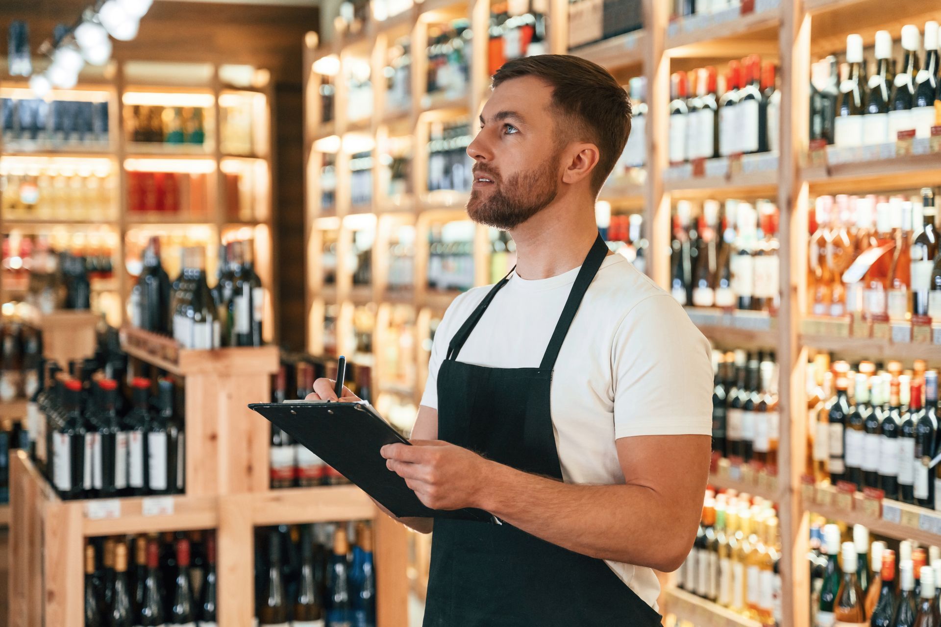 A man in an apron is holding a clipboard in a wine store.