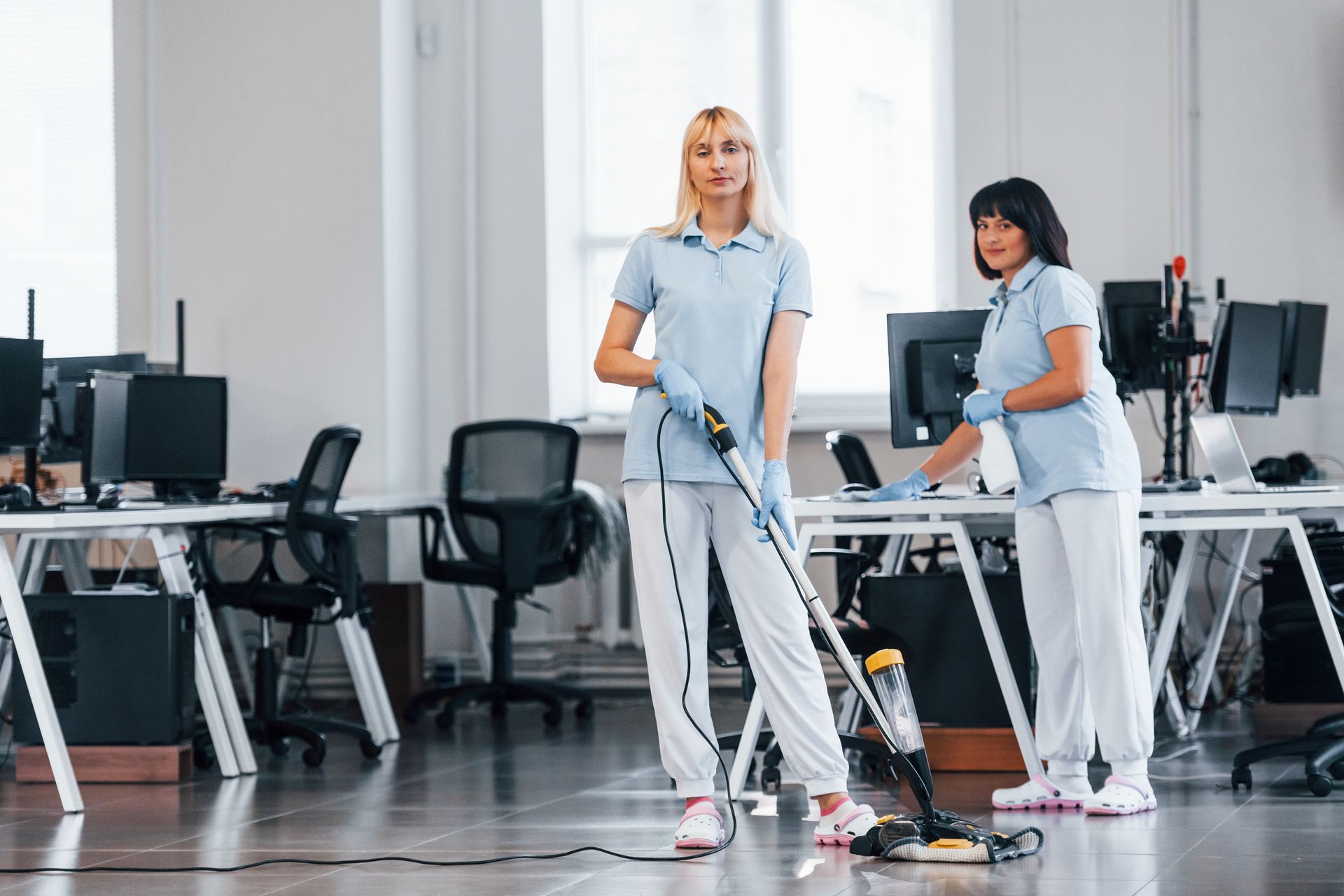 Two women are cleaning an office floor with a vacuum cleaner.